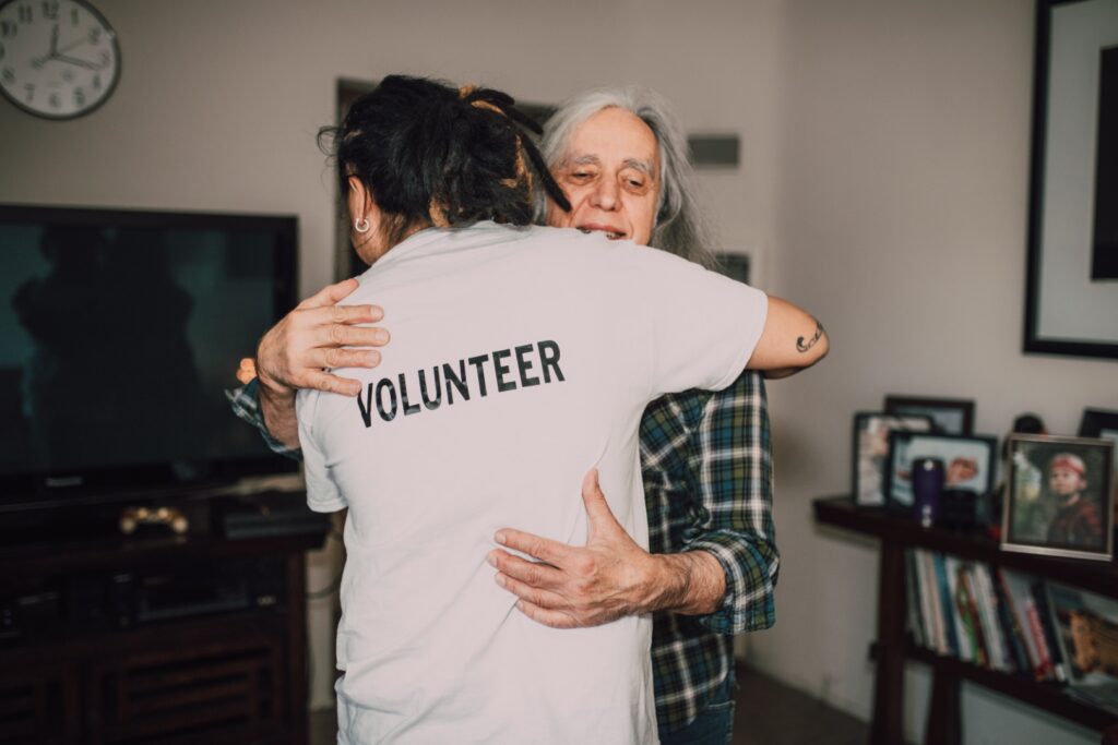 Wolontariusz w koszulce z napisem "VOLUNTEER" przytula seniora. W pokoju są książki, zdjęcia rodzinne, telewizor, zegar ścienny.